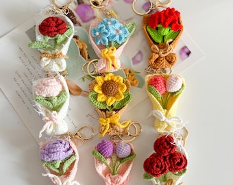 May include: A collection of handmade crochet flower bouquet keychains. Each keychain features a different flower design, including roses, sunflowers, and tulips, in various colors. The bouquets are wrapped in decorative paper and tied with ribbons.