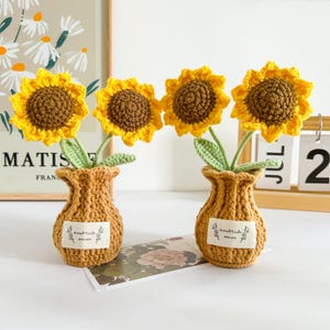 以下が含まれることがあります： 小さな、質感のあるベージュの花瓶に入った、手作りのかぎ針編みのひまわりが4つ。ひまわりの花びらは黄色で、中心は茶色、茎と葉は緑色です。各花瓶には「Handmade with love」と書かれた小さなラベルが付いています。