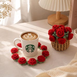 Puede incluir: Una taza de cerámica blanca con el logotipo verde de Starbucks sobre un posavasos redondo de ganchillo color crema, decorado con pequeñas rosas rojas y hojas verdes. Al lado hay una pequeña cesta redonda de ganchillo marrón llena de rosas.