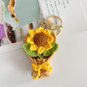 Puede incluir: Llavero de girasol tejido a mano. El llavero presenta un girasol amarillo con centro marrón, hojas verdes y un envoltorio de ramo beige atado con un lazo amarillo. Anillo dorado.
