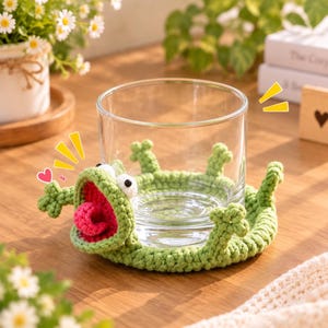 Puede incluir: Un vaso de cristal transparente sobre un posavasos redondo de ganchillo de color verde claro con forma de rana. El posavasos presenta una boca muy abierta con una lengua de color rosa brillante, dos ojos blancos y cuatro patas de rana.