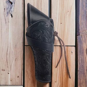 Könnte beinhalten: Schwarze Lederholster mit floralen Verzierungen und weißen Nähten. Die Holster verfügt über eine Gürtelschlaufe und eine Lederschnur. Das Design ist im klassischen Western-Stil gehalten und für eine Schusswaffe geeignet. Die Holster ist auf einem Holzgrund befestigt.