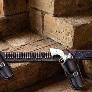 May include: A dark brown leather gun belt featuring two matching holsters, each containing a silver revolver with a cream-coloured grip. The belt includes a series of loops holding metallic cartridges and is secured with a silver-toned metal buckle.