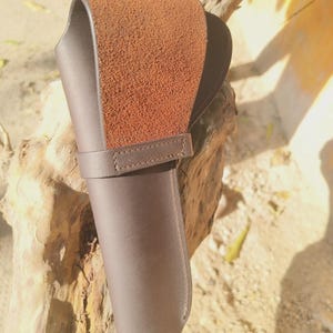 May include: A brown leather knife sheath with a textured, suede-like flap. The sheath has a loop for a belt and a strap to secure the knife. The leather is a rich, dark brown color, and the stitching is visible.