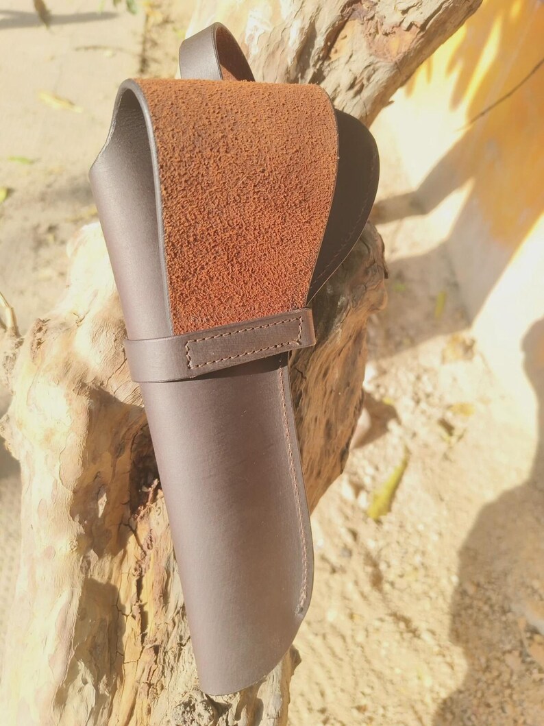 May include: A brown leather knife sheath with a textured, suede-like flap. The sheath has a loop for a belt and a strap to secure the knife. The leather is a rich, dark brown color, and the stitching is visible.
