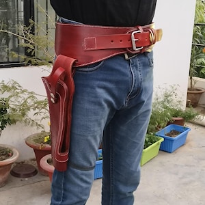 Puede incluir: Un cinturón de pistola de cuero rojo con una funda a juego. El cinturón ancho tiene una hebilla grande y una trabilla más pequeña. La funda está sujeta al cinturón y tiene una correa para la pierna. Costuras blancas.