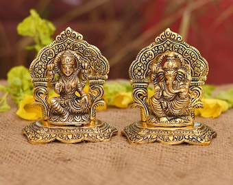 May include: Two gold-colored statues of Hindu deities. The statue on the left depicts a seated female figure, while the one on the right shows a seated elephant-headed figure. Both statues have ornate, arched frames and detailed bases. The statues are on a brown surface.