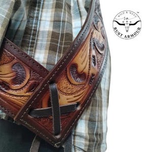 May include: Close-up of a brown leather guitar strap with detailed floral tooling. The strap has a dark brown border and a cross-stitched design. The Wild & West Rust Armour logo is visible.