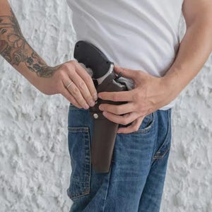 May include: A black handgun is partially visible in a brown leather holster. The holster is attached to a person's waist, secured by a belt loop. The person is wearing blue jeans and a white t-shirt.