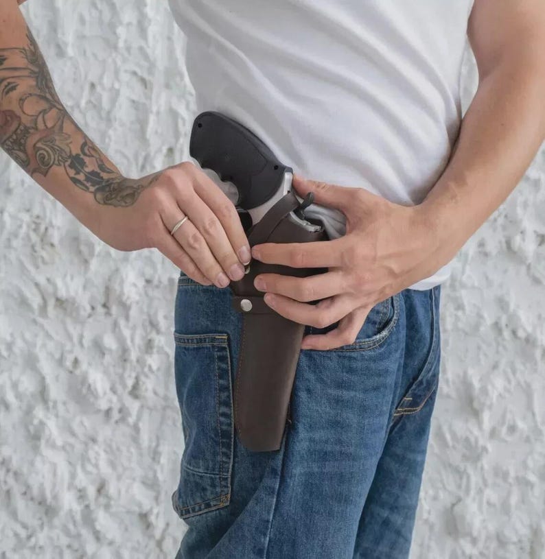May include: A black handgun is partially visible in a brown leather holster. The holster is attached to a person's waist, secured by a belt loop. The person is wearing blue jeans and a white t-shirt.