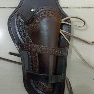 May include: A dark brown leather holster with decorative embossed patterns. The holster features a belt loop, a gun compartment, and leather ties. The leather has a worn appearance, suggesting a vintage or rustic style.