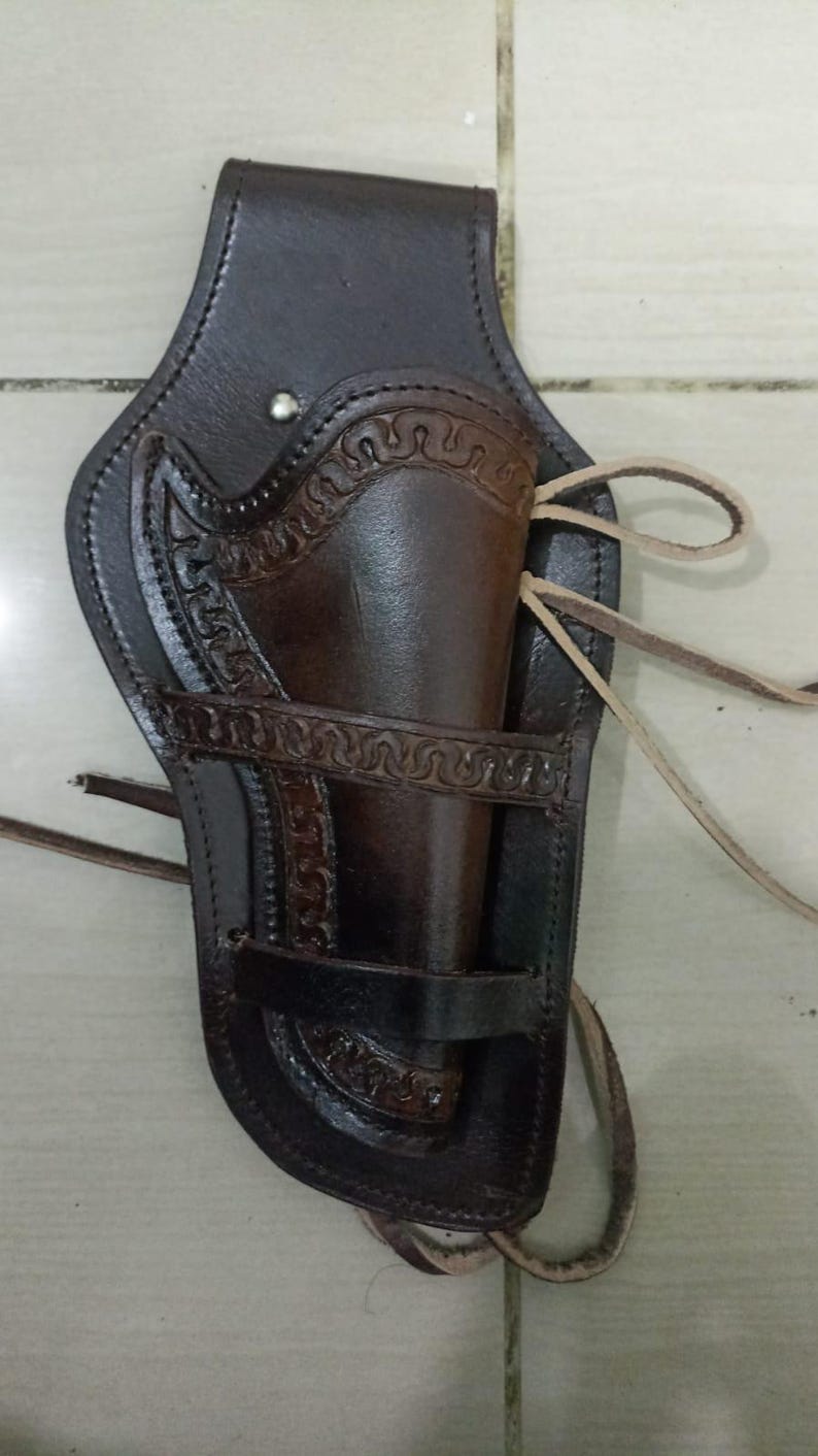 May include: A dark brown leather holster with decorative embossed patterns. The holster features a belt loop, a gun compartment, and leather ties. The leather has a worn appearance, suggesting a vintage or rustic style.