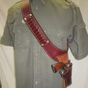 Könnte beinhalten: Ein burgunderfarbenes Schulterholster aus Leder mit passendem Patronengurt, weißen Nähten und silbernen Nieten. Das Holster hält einen Revolver. Die Artikel werden über einem grünen kurzärmeligen Hemd präsentiert.