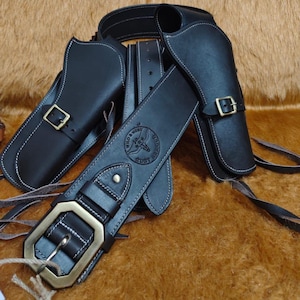 Könnte beinhalten: Ein schwarzes Leder-Waffenholster-Set mit Gürtel und zwei Holstern. Das Set verfügt über messingfarbene Schnallen und weiße Nähte. Der Gürtel hat ein dekoratives Emblem mit dem Text "Wild & West Armour".