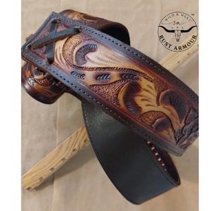May include: A hand-tooled leather guitar strap with detailed floral and scrollwork patterns. The strap has a dark brown edge and a lighter tan central section, with a black underside. The "RUST ARMOUR" logo is visible.
