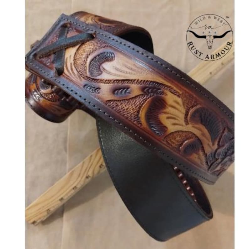 May include: A hand-tooled leather guitar strap with detailed floral and scrollwork patterns. The strap has a dark brown edge and a lighter tan central section, with a black underside. The "RUST ARMOUR" logo is visible.
