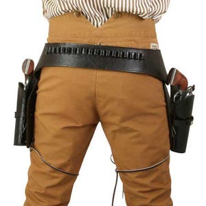 Puede incluir: Un cinturón de pistola de cuero negro con presillas para balas y dos fundas, cada una con un revólver. El cinturón se lleva sobre pantalones marrones. La persona lleva una camisa a rayas.