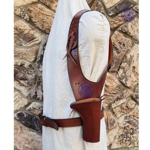 Puede incluir: Una funda de hombro de cuero marrón con cinturón a juego. La funda tiene una bolsa para una pistola y se asegura con correas y una hebilla. Un emblema púrpura es visible en la funda.