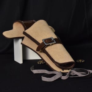 May include: A beige leather holster with brown edging and a buckle fastening. The holster is designed to hold a small item and has a strap. The background is black.