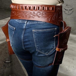 May include: Brown leather gun belt with floral tooling and cartridge loops. The belt is worn with a matching holster and leg tie. The image shows the belt and holster against a background of blue jeans.