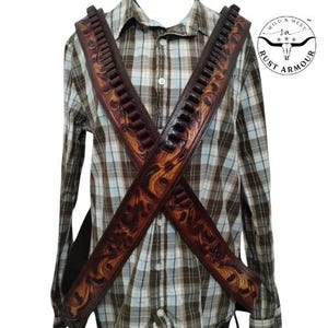 May include: Brown leather gun belt with decorative floral tooling, worn over a plaid shirt. The belt features loops for ammunition and is designed in a cross-body style. The Rust Armour logo is visible in the upper right corner.