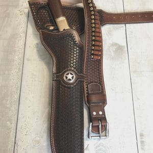 May include: A dark brown leather holster with a basket-weave pattern and a silver star concho, containing a lever-action rifle with a wooden stock and brass receiver. An attached leather belt features loops holding twelve brass-cased cartridges.