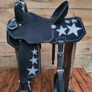 May include: A black western saddle featuring silver glitter star appliques and silver stud detailing along the edges. The saddle includes a horn, a seat, stirrups, and leather straps, with a prominent star design on the skirt and fender.