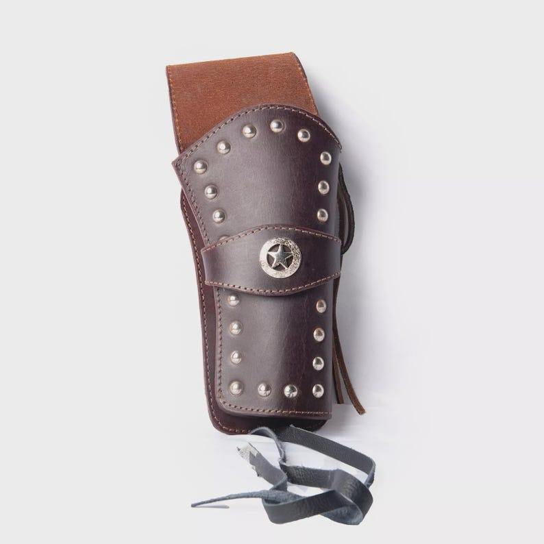 May include: A brown leather holster with silver studs and a star-shaped emblem. The holster has a belt loop and a strap. The design is reminiscent of the Wild West. The holster is made of leather.