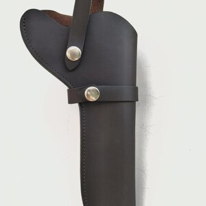 May include: Black leather holster with a loop handle and a strap secured by silver-colored snaps. The holster is designed to hold a firearm and features a stitched edge.