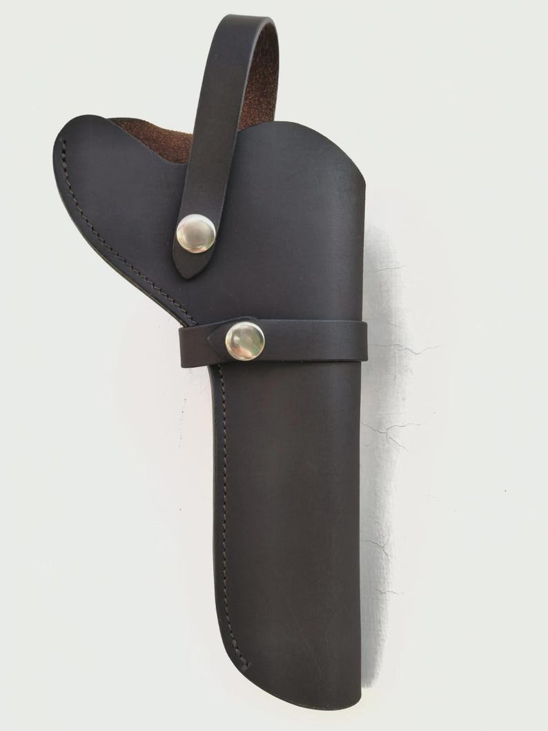May include: Black leather holster with a loop handle and a strap secured by silver-colored snaps. The holster is designed to hold a firearm and features a stitched edge.
