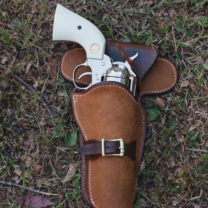 May include: A silver revolver with a cream-colored handle is partially holstered in a brown leather holster. The holster has a belt loop and a buckle. The image is taken outdoors on a bed of grass and leaves.