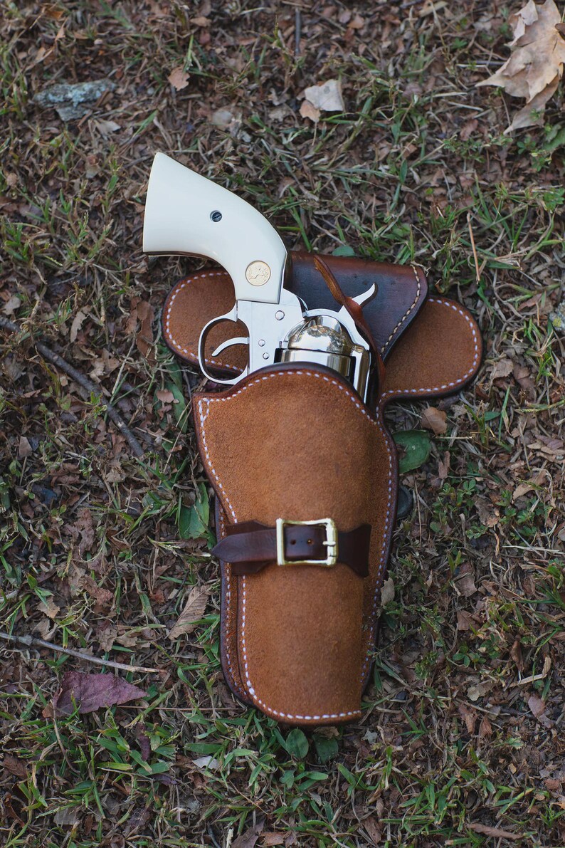 May include: A silver revolver with a cream-colored handle is partially holstered in a brown leather holster. The holster has a belt loop and a buckle. The image is taken outdoors on a bed of grass and leaves.