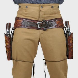 Puede incluir: Un cinturón de cuero marrón con dos fundas, cada una con un revólver. El cinturón tiene una hebilla de cuero marrón y adornos decorativos. Las fundas también son de cuero marrón con adornos decorativos. La persona lleva pantalones color caqui.