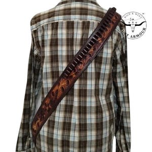 May include: A brown leather gun belt with decorative floral tooling and cartridge loops. The belt is worn over a plaid shirt. The Rust Armour logo is visible in the upper right corner. The belt is a western-style accessory.
