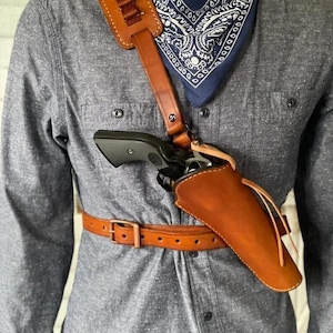 Puede incluir: Una funda de hombro de cuero marrón con un cinturón a juego. La funda contiene un revólver negro. Una bandolera con bucles de bala marrones está unida a la correa del hombro. Un pañuelo azul con estampado de cachemira se lleva alrededor del cuello.