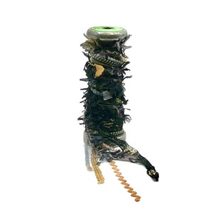 May include: A spool of decorative ribbon with a metallic green top and a base. The ribbon is wrapped with dark green, black, and gold fabric and trim. Several strands of beige and brown ribbon are unwound from the spool.