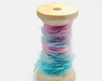 May include: A wooden spool wrapped with fuzzy yarn in a gradient of pastel colors, transitioning from light pink at the top to a vibrant turquoise blue at the bottom. The yarn has a soft, textured appearance with loose fibers visible throughout.