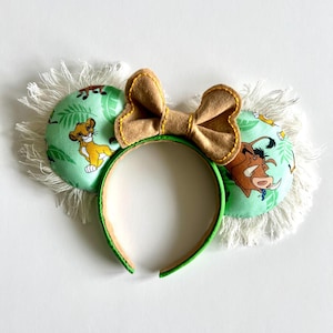 May include: Green headband with a brown bow and two fabric ears. The ears are decorated with a green jungle print featuring Simba, Pumbaa, and Timon from Disney's The Lion King. The ears have white fringe around the edges.