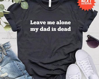 Könnte beinhalten: Schwarzes T-Shirt mit dem weißen Text "Leave me alone my dad is dead". Das Bild zeigt auch blaue Jeans, weiße Turnschuhe und eine Sonnenbrille. Das T-Shirt ist aus einem weichen Material.