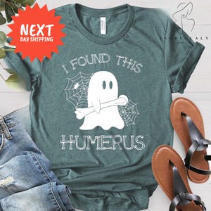 May include: A teal t-shirt with a white graphic of a ghost holding a bone, with the text "I FOUND THIS HUMERUS". The design includes spiderwebs. The shirt is paired with blue jeans and brown sandals.