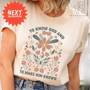 May include: Cream-colored t-shirt with a floral design and the text "TO KNOW GOD AND TO MAKE HIM KNOWN." The design features a large central flower in muted tones, surrounded by smaller flowers and leaves. The shirt has a round neckline and short sleeves.