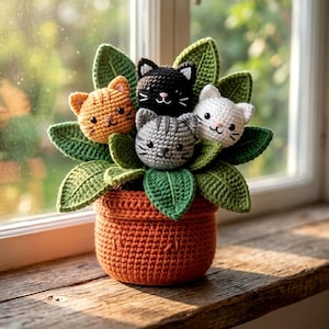 May include: A crocheted cat planter featuring four cat faces amongst green leaves. The cats are orange, black, white, and grey. The orange planter sits on a wooden surface near a window.