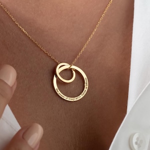 Könnte beinhalten: Eine goldene Halskette mit einem Doppelkreis-Anhänger. Die Kreise sind ineinander verschlungen und haben ein zartes, graviertes Design.