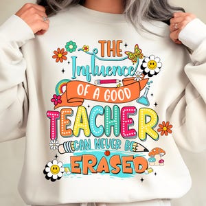 May include: A cream-coloured sweatshirt featuring a vibrant graphic. The text reads "THE Influence OF A GOOD TEACHER CAN NEVER BE ERASED." The design incorporates flowers, a rainbow, a butterfly, and pencils.