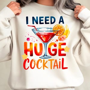 May include: White sweatshirt with the text "I NEED A HUGE COCKTAIL" in a colorful, watercolor-style design. The graphic includes a cocktail glass with a red drink, an orange slice, and star accents.