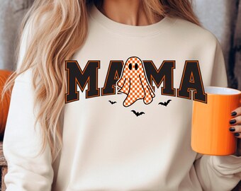 May include: Cream-colored sweatshirt with the word "MAMA" in black block letters with orange trim. A checkered orange and white ghost graphic is in the center, with three small black bats below. An orange mug is held in the hand.