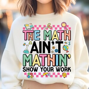May include: White sweatshirt with the text "THE MATH AIN'T MATHIN' SHOW YOUR WORK" in colorful, dotted letters. The design includes math-related graphics like scissors, a magnifying glass, and a globe. The sweatshirt is a casual, comfortable style.