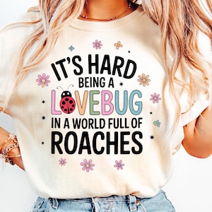 May include: Cream-colored t-shirt with the text "IT'S HARD BEING A LOVEBUG IN A WORLD FULL OF ROACHES" in a colorful, whimsical font. The design includes a ladybug and floral accents.
