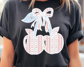 May include: Dark gray t-shirt featuring a whimsical cherry design. The cherries are adorned with a white lace trim and a bow with a blue gingham pattern. The cherry design has a red and white pattern.