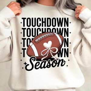 Puede incluir: Sudadera color crema con un gráfico de fútbol americano. El balón es marrón con rayas blancas y un diseño en forma de corazón. Las palabras "TOUCHDOWN" y "Season" están impresas en negro.